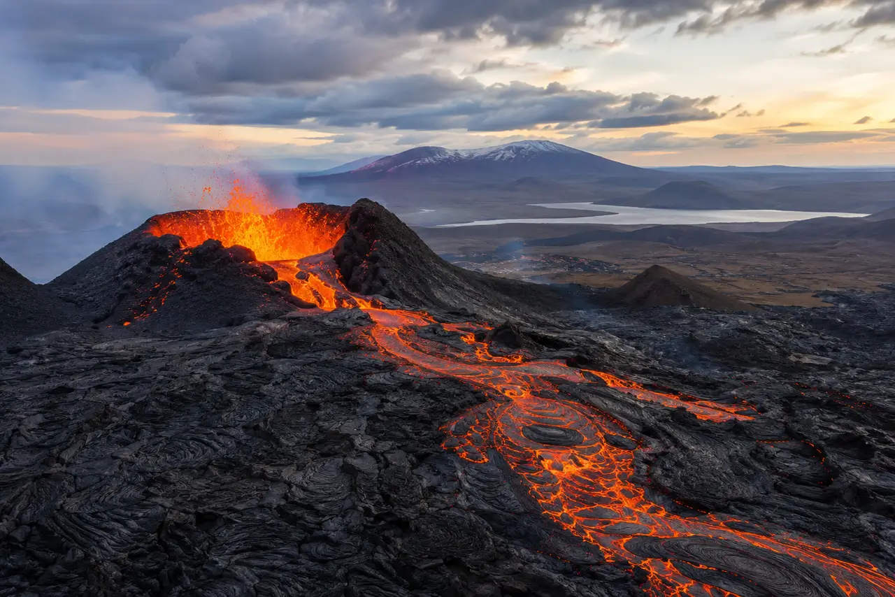 Erupción volcánica con lava fluyendo sobre terreno rocoso y paisaje geológico amplio al fondo