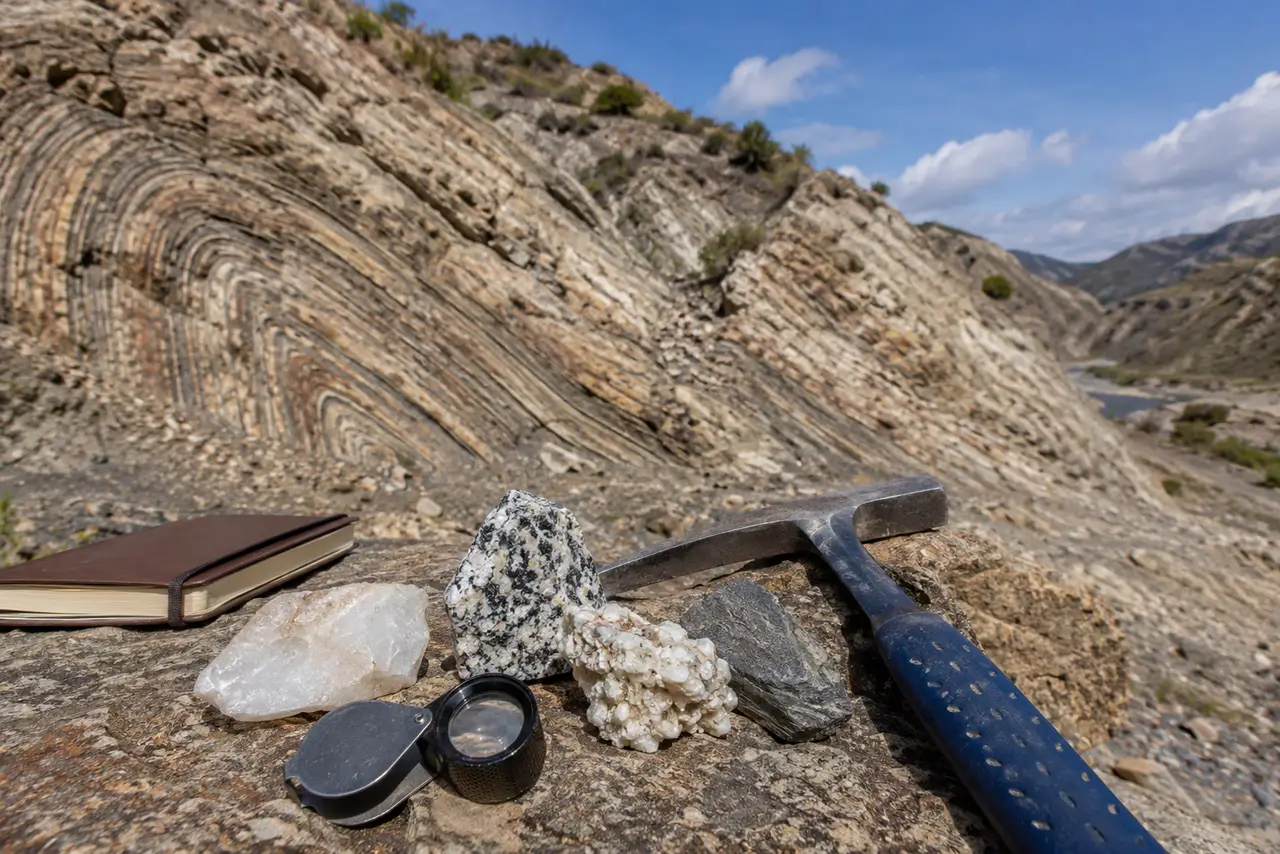 Afloramiento rocoso plegado con martillo geológico, lupa y muestras minerales en un paisaje claro de campo